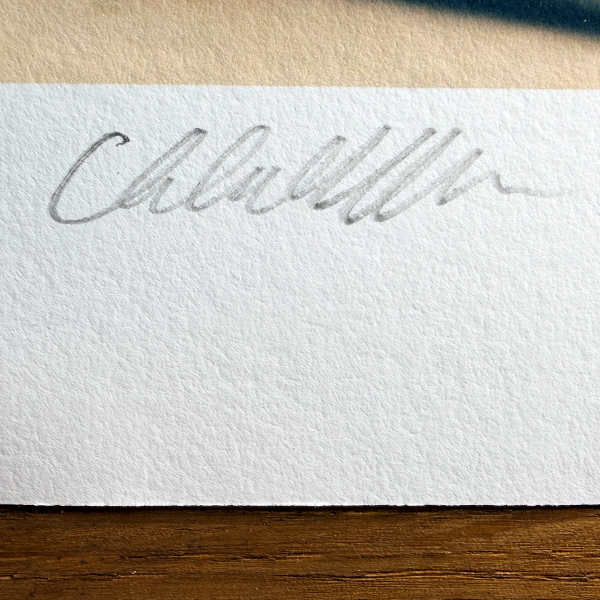 Close-up of Chloe Albert's hand signature and edition number in pencil on the deckled border of a Hahnemühle fine art print, lit with raking light to show the paper texture.