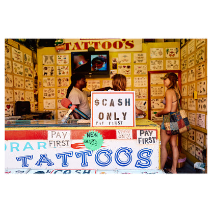 The interior of a Coney Island tattoo parlor, its walls covered in flash art, a hand-painted '$ Cash Only — Pay First' sign in the centre. A color fine-art photograph by Chloe Albert from the Coney Island series, printed on Hahnemühle Photo Rag Bright White.