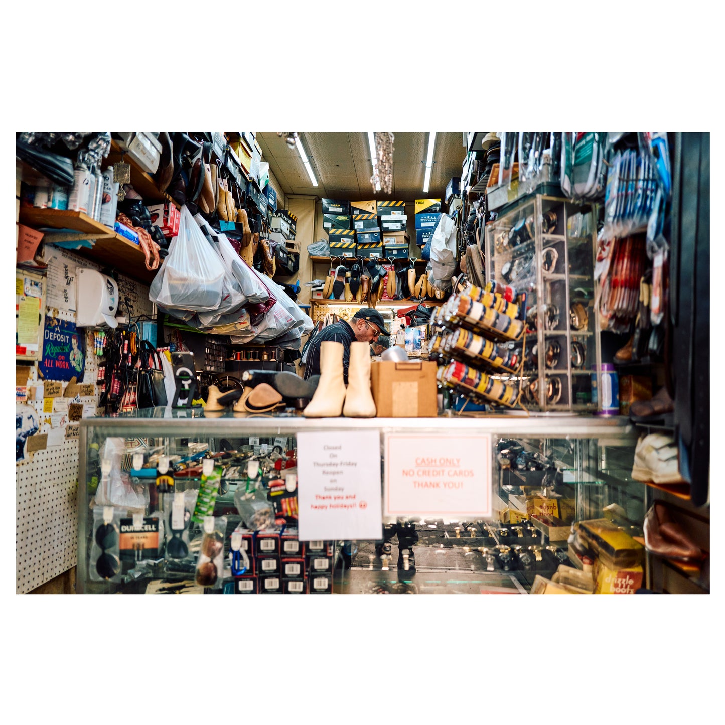 The interior of a cobbler's shop in Chelsea, New York, shelves lined with shoes and boots and the cobbler at work behind the counter. A color fine-art photograph by Chloe Albert, printed on Hahnemühle Photo Rag Bright White fine art paper.