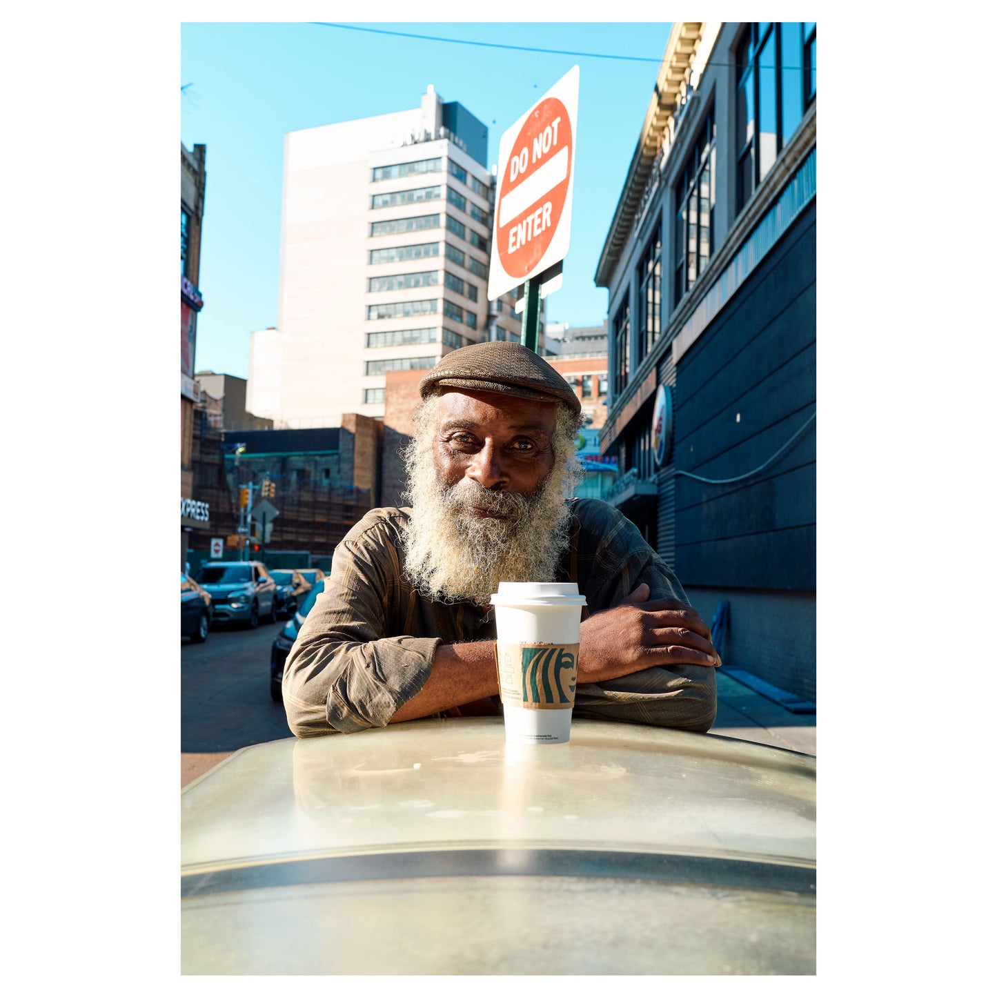 An older man in a newsboy cap leans on a round metal table with a paper coffee cup, a red 'Do Not Enter' sign beside him on Jay Street, Brooklyn, New York. A color fine-art photograph by Chloe Albert, printed on Hahnemühle Photo Rag Bright White fine art paper.