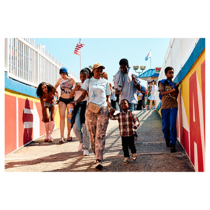 A crowd walks down a ramp between boldly painted red and yellow walls at the Coney Island boardwalk, New York. A color fine-art photograph by Chloe Albert from the Coney Island series, printed on Hahnemühle Photo Rag Bright White.