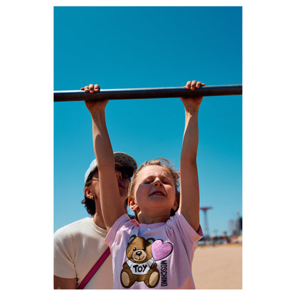 A child hangs by both hands from a boardwalk railing against a clear blue sky at Coney Island, feet lifted off the ground. A color fine-art photograph by Chloe Albert from the Coney Island series, printed on Hahnemühle Photo Rag Bright White.