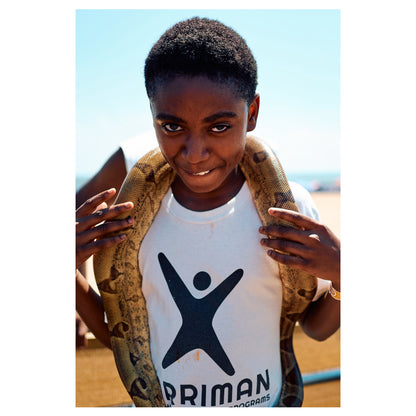 A young person in a white T-shirt stands on the Coney Island boardwalk with a large python draped across their shoulders, the beach behind. A color fine-art photograph by Chloe Albert from the Coney Island series, printed on Hahnemühle Photo Rag Bright White.