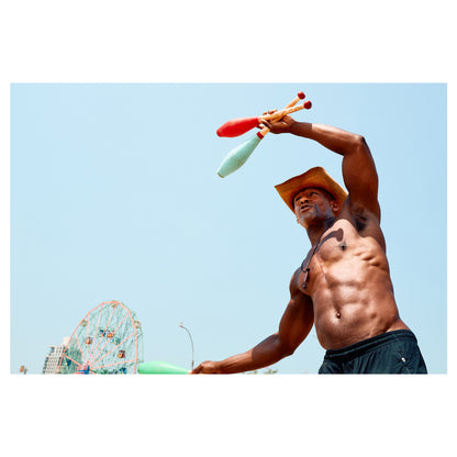 A shirtless man in a straw cowboy hat juggles clubs on the Coney Island boardwalk, amusement rides and blue sky behind him. A color fine-art photograph by Chloe Albert from the Coney Island series, printed on Hahnemühle Photo Rag Bright White.