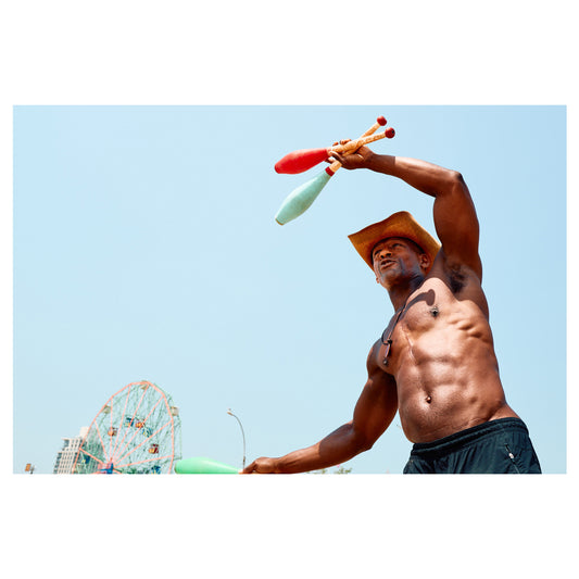 A shirtless man in a straw cowboy hat juggles clubs on the Coney Island boardwalk, amusement rides and blue sky behind him. A color fine-art photograph by Chloe Albert from the Coney Island series, printed on Hahnemühle Photo Rag Bright White.