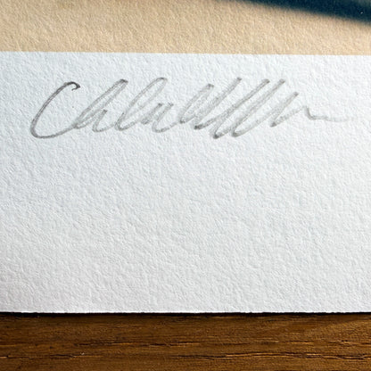 Close-up of Chloe Albert's hand signature and edition number in pencil on the deckled border of a Hahnemühle fine art print, lit with raking light to show the paper texture.