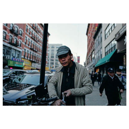 A delivery driver in a flat cap pauses on the move on a Chinatown street in New York, scaffolding and traffic around him. A color fine-art photograph by Chloe Albert, printed on Hahnemühle Photo Rag Bright White fine art paper.